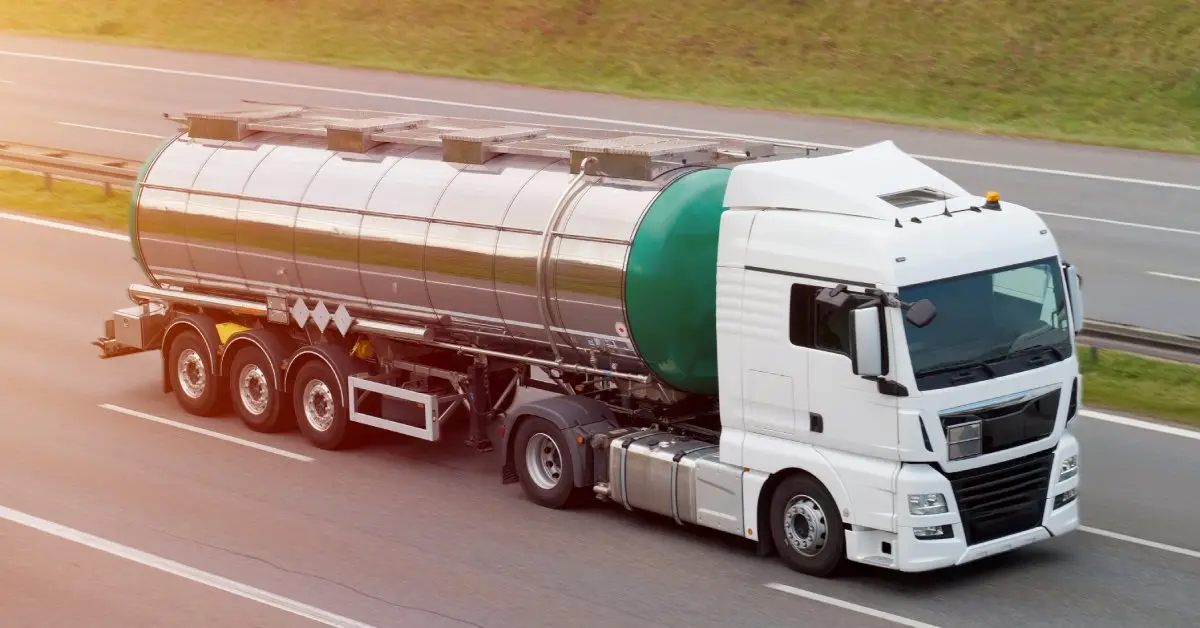 A green and chrome tanker trailer with a white cab and several wheels drives down the road next to the grass.
