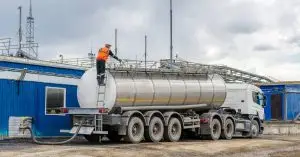 A stainless steel tanker truck is parked in front of a blue building and a worker in an orange vest works on the vehicle.