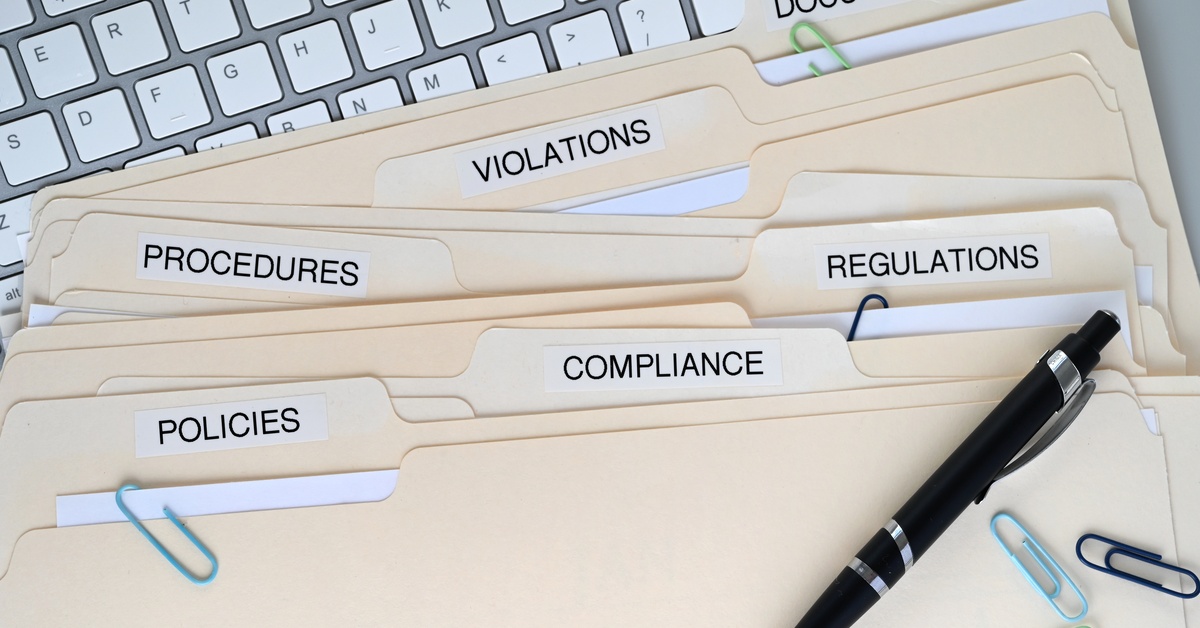 A stack of file folders on a desk labeled: "policies, compliance, regulations, procedures, violations, and documentation."