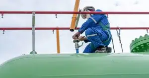 Closeup of a worker in coveralls and personal protective equipment working on a valve on top of a green tanker.